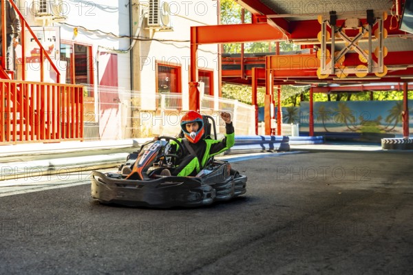Go kart driver in helmet and racing suit crossing finish line on outdoor track, celebrating victory with raised arms after fast, competitive race on sunny summer day