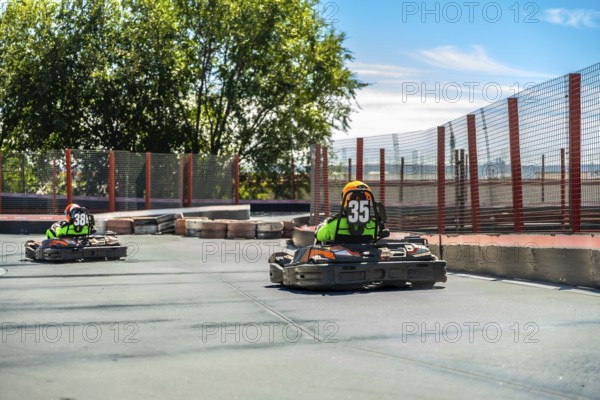 Two competitive go karts with drivers wearing helmets and number vests driving quickly on an asphalt racing circuit, engaging in a thrilling motorsport activity under clear blue skies