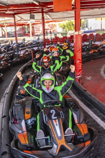 Group of excited people wearing helmets and racing suits, sitting in go karts and raising their fists, lining up on starting grid for a fun race at a karting track