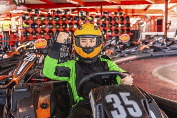 Asian man in racing suit and yellow helmet sits in a go kart on track, smiling and pumping fist in excitement celebrating a win or gearing up for a fast, adrenaline filled race