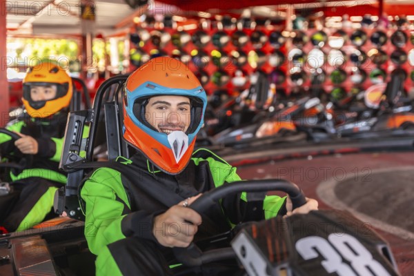 Young man wearing a colorful helmet and racing suit, smiling and driving a go kart on a track with other karts and a helmet wall in the background, conveying excitement and fun