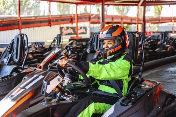 Young woman in a racing suit and helmet sitting in her go kart at the starting line, smiling and anticipating the fun and friendly competition on the circuit