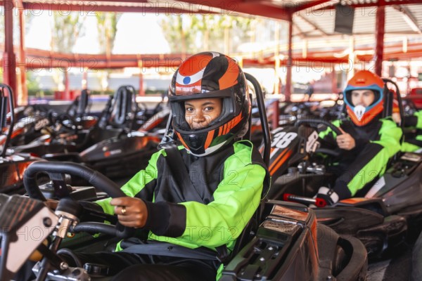 Young woman smiling in a black and neon green racing suit and orange helmet, sitting in a go kart on a track with another driver in the background, preparing for a fun karting race