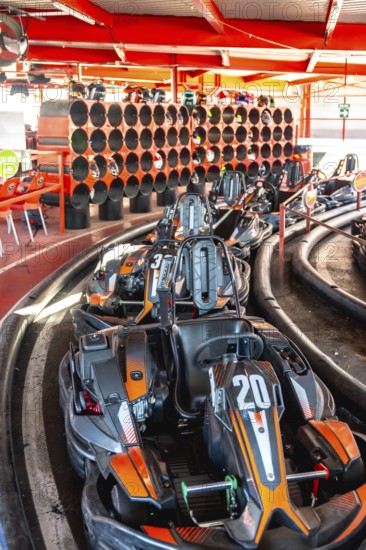 Go karts with orange and black details resting in a queue at the starting line of an indoor track, with helmets stored on a rack in the background, preparing for a race