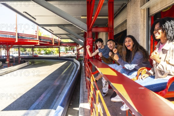 Diverse group of young friends is enthusiastically cheering and watching a go kart race from an elevated platform, enjoying leisure time and an exciting activity together at the track