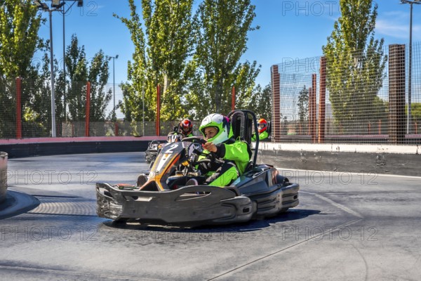 Drivers are speeding go karts around a curve on an outdoor asphalt track, wearing protective helmets and bright racing suits, enjoying the thrill of competition and motor sports on a sunny day
