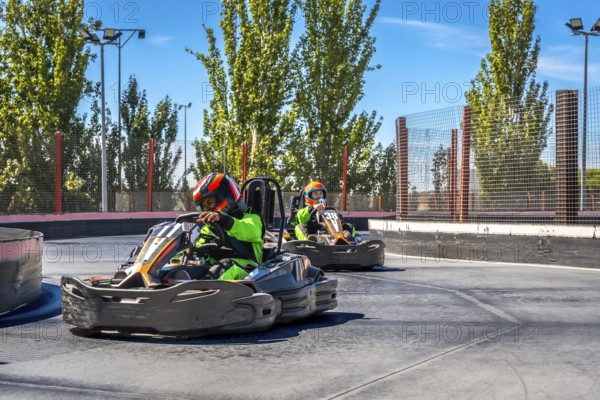 Two children in helmets and protective gear race go karts on an outdoor track, enjoying the speed, competition and thrill of karting on a bright day