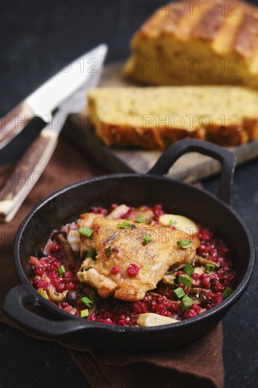 Roasted chicken thigh in cast iron skillet with lingonberry sauce. Slices of fresh bread are placed beside the pan on a wooden board. A knife and fork are shown