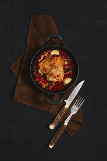 A chicken dish is served in a black cast iron pot. The pot contains spicy lingonberry sauce. Two utensils are placed beside the pot on a brown cloth. The scene is set on a dark surface