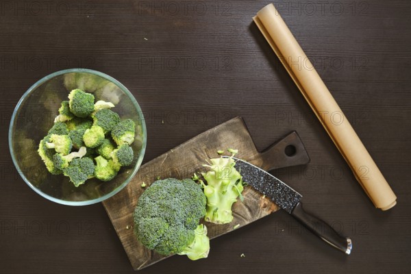 Broccoli is being cut on a wooden board. A bowl of chopped broccoli sits nearby. The kitchen is organized and ready for the cheese to be added to the smashed broccoli