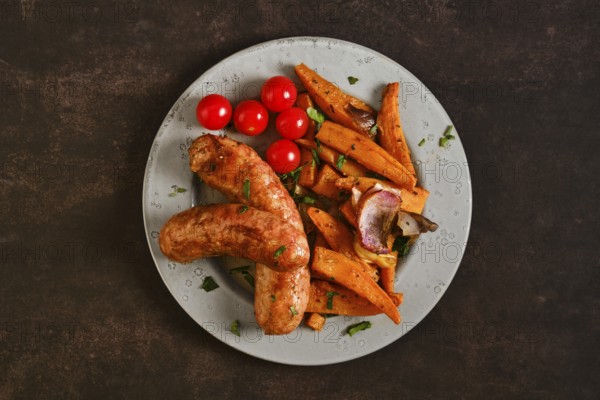 Top view of natural casing sausages on a plate alongside roasted sweet potatoes, onions, and cherry tomatoes. The meal shows a mix of colors with simple sides on a dark background