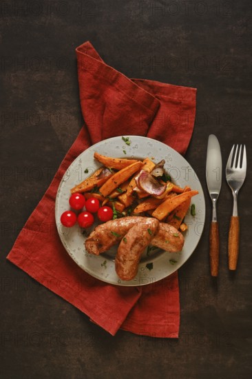 Natural casing sausages are placed on a green plate alongside roasted sweet potatoes and cherry tomatoes. A red napkin and utensils are arranged on a dark wooden table under soft light