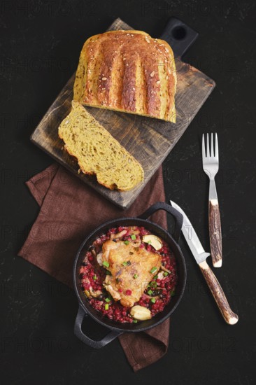 Top view of roasted chicken thigh with lingonberry in a pan on a brown cloth. Freshly baked bread sits on a wooden board beside the dish. A fork and knife are ready for serving