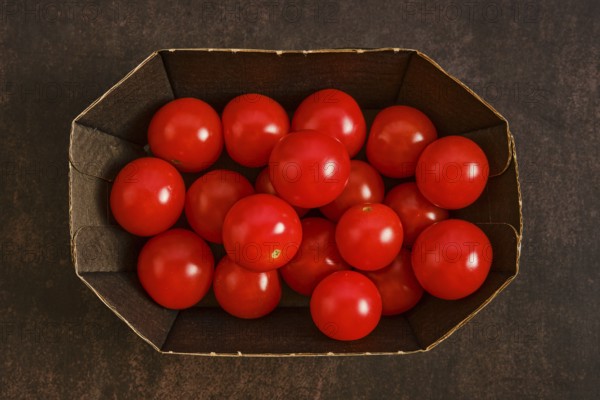 Cardboard box holds many red cherry tomatoes. The tomatoes are bright and round, placed together without any leaves or stems. The background is dark, making the colors stand out
