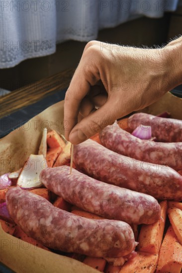 A hand is placing a toothpick into natural casing sausages that rest on a bed of sweet potatoes and onions. The background shows a simple kitchen with soft lighting in the late afternoon