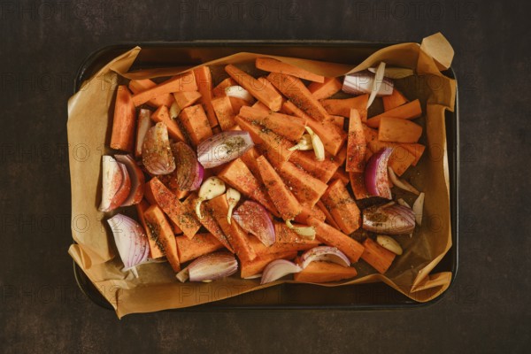 Top view of sweet potatoes, red onion and garlic seasoned on parchment lined tray, prepared for roasting, representing healthy home cooking and plant based lifestyle