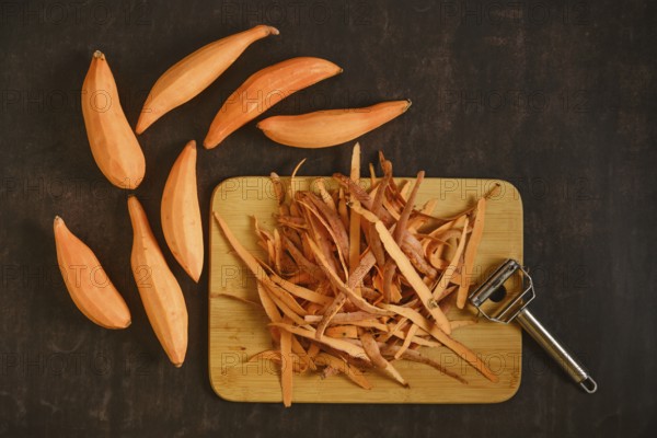 Peeled sweet potatoes with a vegetable peeler on a wooden cutting board. The skin is piled next to several whole sweet potatoes. The kitchen is tidy and organized