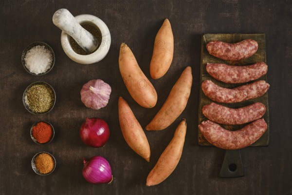 Natural casing sausages are placed next to sweet potatoes and several bowls of spices on a wooden table. All ingredients are arranged neatly with garlic and red onions in view