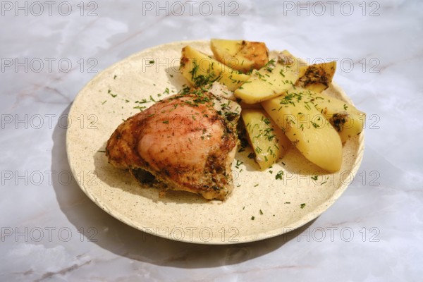 A portion of baked chicken is placed on a plate alongside cut potatoes. The chicken has a bit of herbs sprinkled on top for flavor. The dish is on a table with a light surface
