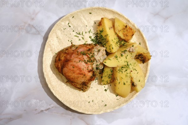 A chicken thigh is placed beside several pieces of seasoned potatoes on a round plate. The dish has a sprinkle of herbs on top. The setting is simple and focuses on the food