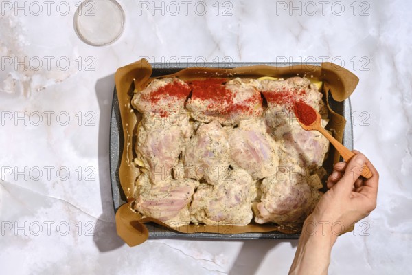 A person spreads smoked paprika over chicken thighs placed in a baking tray lined with parchment paper. The kitchen is well lit and organized, with a few items in the background