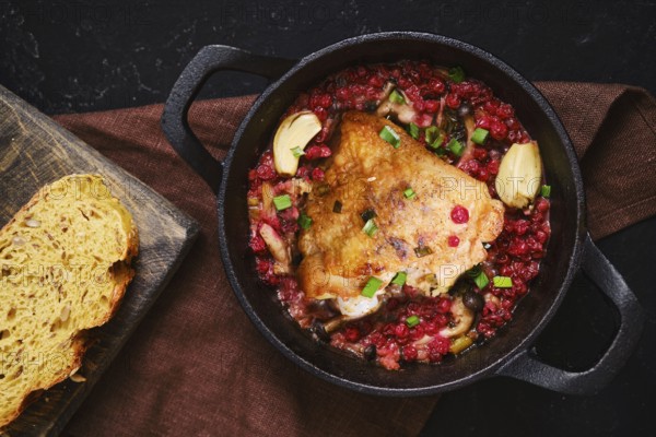A chicken piece is cooking in a pot filled with lingonberry and garlic. A slice of bread is placed on a wooden board nearby. The setting looks like a kitchen countertop
