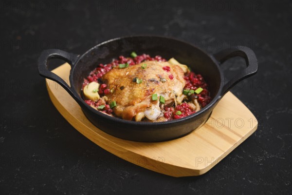 A cast iron skillet holds a serving of cooked chicken topped with lingonberry and chopped spring onion. The dish is placed on a wooden board. The setting is dark, creating focus on the food