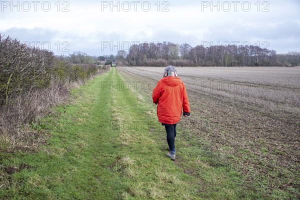 Person wearing bright orange coat walking along grassy path next to hedgerow and winter field, Sutton, Suffolk, England, UK