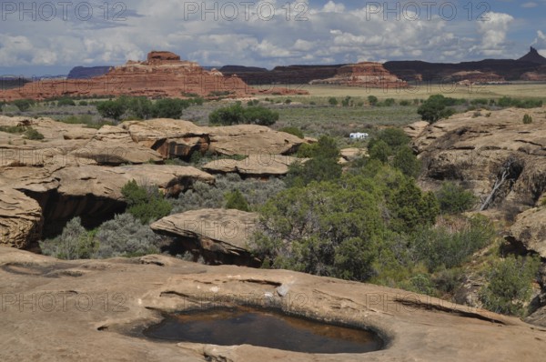 Wide rocky landscape with natural water basins and large red rocks in the background under a cloud cover, Canyonlands National Park, Utah, USA