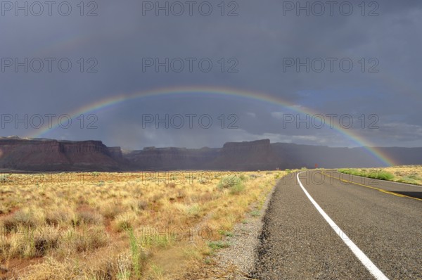 Another rainbow over a long, empty road in a colorful desert landscape with cloudy sky, Canyonlands National Park, Utah, USA