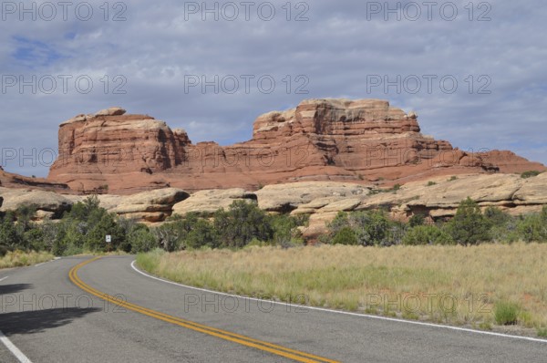 A winding country road runs in front of an impressive red rock formation under cloudy skies, Canyonlands National Park, Utah, USA