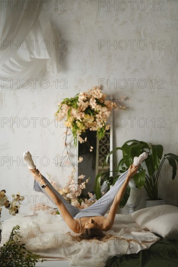 A woman is lying on a bed and stretching her legs in the air. The room has flowers and green plants. Soft light enters from nearby windows, creating a warm atmosphere during the day