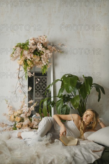 A woman relaxes on a bed while reading a book in a sunny room. She is surrounded by plants and flowers. There is a mirror in the background adding depth to the space