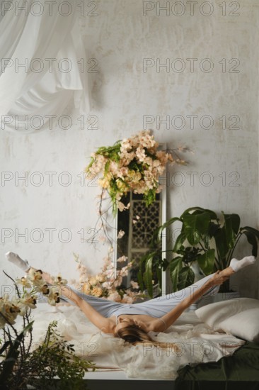 Woman in pajamas stretches on a bed in a bright room filled with plants and flowers. The stretching exercise shows the person's flexibility and focus