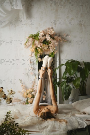 A woman is lying on her back while stretching her legs up in the air. The room has many plants and floral decorations. Soft light comes through the window and bathes the space