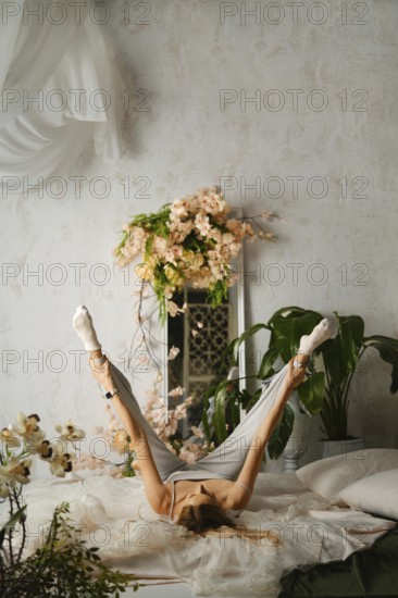 Flexible woman lies on her back with her legs up in the air on a bed. The room is filled with flowers and plants, creating a lively atmosphere. Natural light enters from a window, enhancing the scene