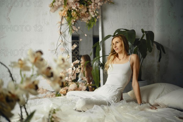 A woman sits on a bed in a bedroom decorated with flowers and plants. Natural light fills the space, adding to the relaxed mood of the moment