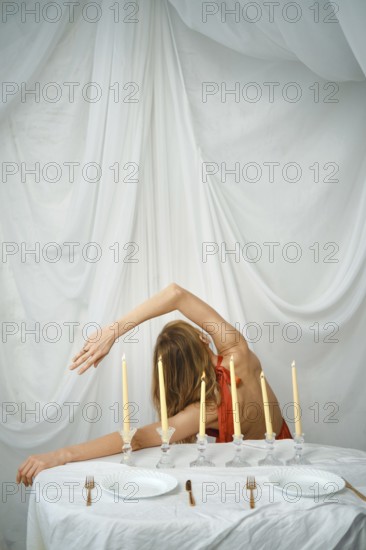 A woman is seen bending gracefully at a table set with candles. The table has white plates and forks, and the backdrop features soft white fabric. It is evening and the scene is set indoors