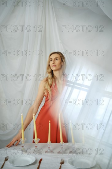 A woman stands near a table that has candles, plates, and cutlery. The soft light comes through the drapes. The setting looks bright and airy. She poses with a thoughtful expression
