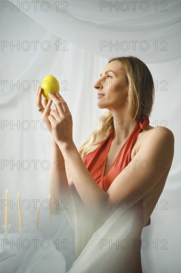 A woman stands indoors, holding a lemon in her hands. She gazes at it with focus. Soft light filters through a draped backdrop. Candles sit nearby, adding to the quiet atmosphere