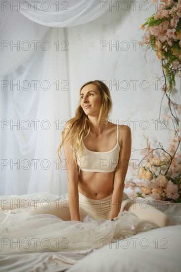 Pleased woman sits on a bed wearing light clothing. She has long hair and smiles while looking away. Flowers decorate the area, making it bright and inviting. Sunlight fills the room and highlights her