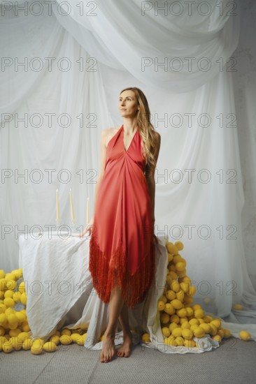 Barefoot woman poses near a table covered with a white cloth. Lemons surround the table as candles are lit. The colors stand out in the bright space. She appears thoughtful and relaxed in her red dress