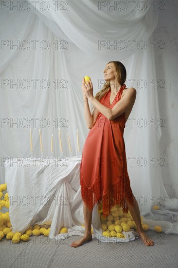 Woman stands in a studio holding a lemon while facing a table decorated with candles and surrounded by lemons. The background features draped fabric and the setting is bright and airy