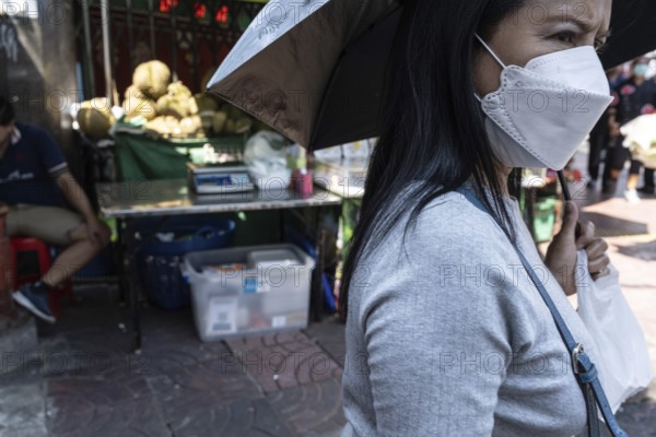Bangkok, Thailand. March 25th 2025. A local woman with an umbrella and wearing a mask due to excessive heat and air pollution in the hot summer climate of Bangkok, Thailand
