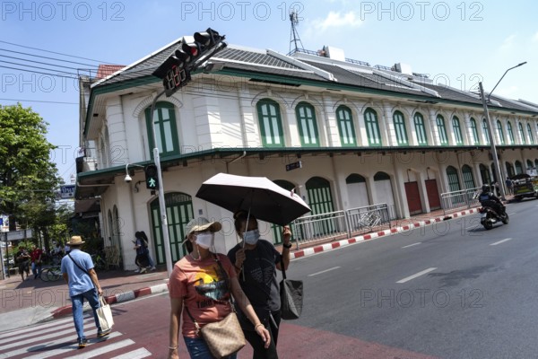 Bangkok, Thailand. March 25th 2025. People crossing the street with masks and an umbrella due to poor air quality and excessive summer heat near Wat Mangkon metro station, Bangkok, Thailand