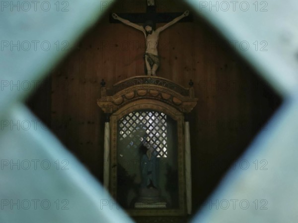Wooden statue of Jesus on the cross through a grid in a dark chapel, hiking in the Frankenwald nature park Park, Upper Franconia