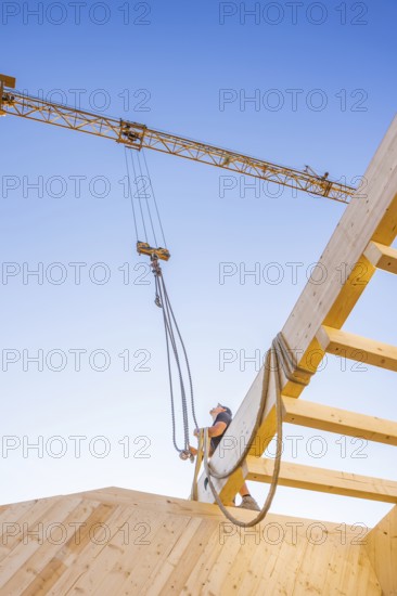 Worker handles a rope next to a crane under a blue sky on a construction site, timber house construction, Gechingen, Germany