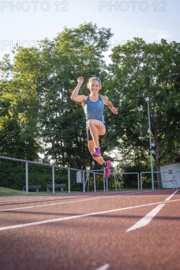 A person wearing blue sportswear jumps with raised arm on the running track in sunshine, middle distance runner Katharina Jaiser, Gechingen, Germany