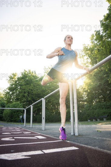 Woman stretching on a track in sunlight, holding onto the railing, surrounded by trees, middle distance runner Katharina Jaiser, Gechingen, Germany