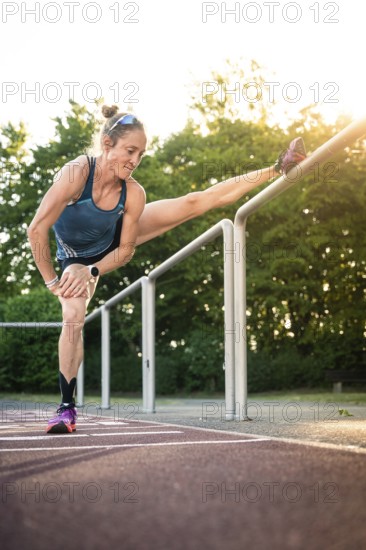 Woman stretches leg on railings in sunshine surrounded by trees near park, middle distance runner Katharina Jaiser, Gechingen, Germany
